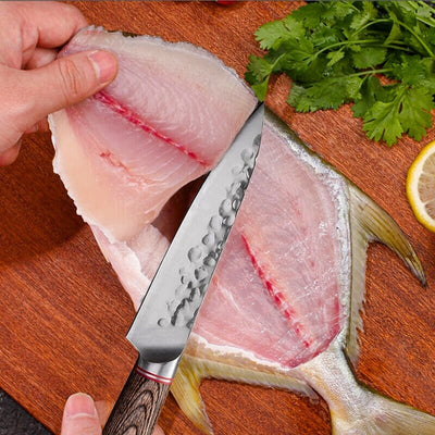 Personne en train de couper un poisson avec un couteau de cuisine avec un manche en bois sur une planche à découper en bois, à côté du persil et une tranche de citron.