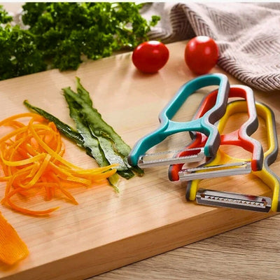 3 économes, 1 de couleur bleue et noire, 1 autre de couleur rouge et noire et 1 dernier de couleur orange et noire posés l'un sur l'autre sur une planche à découper en bois, à côté des légumes coupés en lamelles et en julienne.