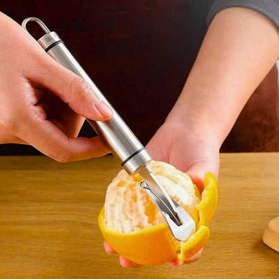 Personne en train d'éplucher une orange avec un éplucheur adapté, elle tient l'orange de la main gauche au-dessus d'une table et l'éplucheur dans la main droite, il enlève morceaux de peau par morceaux de peau.