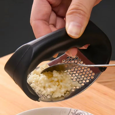 Presse ail tenu par une personne et qui enlève, à l'aide d'une cuillère, l'ail pressé se trouvant sur le presse ail pour le mettre dans une assiette.