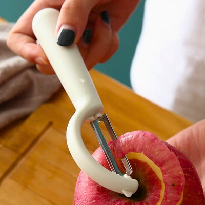 Personne en train d'éplucher une pomme avec un économe blanc au-dessus d'une planche à découper en bois, l'épluchure passe dans la lame de l'appareil.