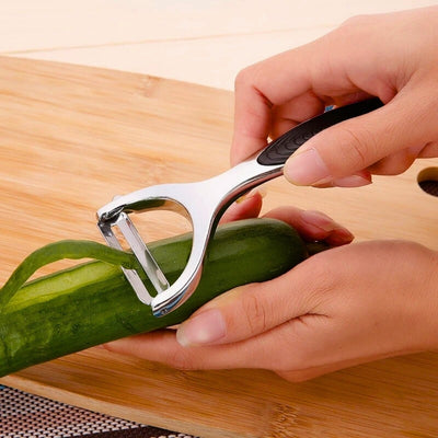 Personne en train d'éplucher un concombre avec un éplucheur à lame horizontale, elle tient le concombre à l'intérieur de sa main au-dessus d'une découper en bois et l'éplucheur avec sa main droite, un morceau d'épluchures sort de l'appareil.