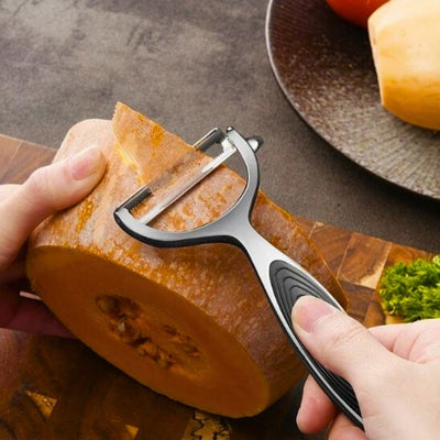 Personne en train d'éplucher une courge avec un éplucheur à lame horizontale, elle tient un morceau de courge avec sa main gauche sur une planche à découper en bois et l'éplucheur avec sa main droite, un morceau d'épluchures sort de l'appareil, à l'arrière une assiette avec des légumes.