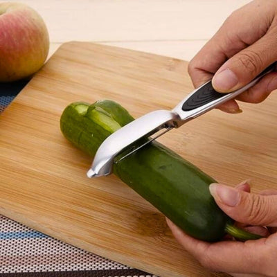 Personne en train d'éplucher un concombre avec un éplucheur à lame verticale, elle tient le concombre avec sa main gauche sur une planche à découper en bois et l'éplucheur avec sa main droite, un morceau d'épluchures sort de l'appareil, à l'arrière une pomme.