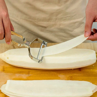 Personne en train de couper une aubergine en lamelle sur une planche à découper en bois, des lamelles passent dans l'économe.