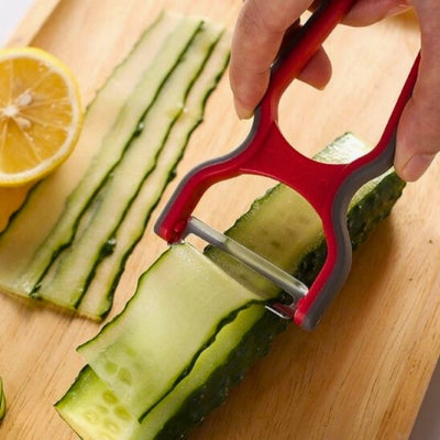 Mains d'une personne en train de couper en lamelle un concombre à l'aide d'un économe de couleur rouge et noire. Elle tient l'économe de la main droite alors que le concombre est posé sur une planche.