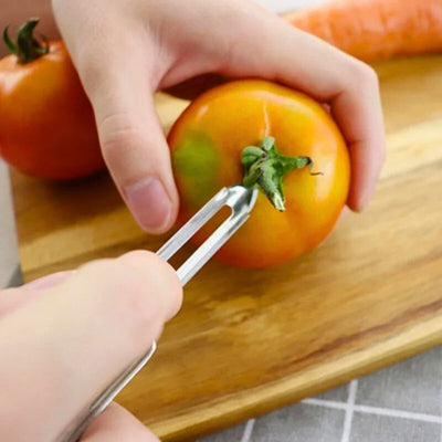 Personne en train de retirer le calice d'une tomate avec le bout de l'éplucheur. La tomate est posée sur une planche à découper et tenu par la main gauche.