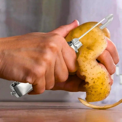 Personne en train d'éplucher une pomme de terre, elle tient dans sa main gauche au-dessus d'une table ne pomme de terre et dans sa main droite l'éplucheur dans lequel pass une épluchure de pomme de terre.
