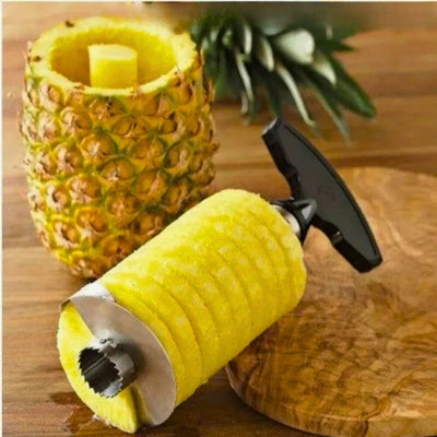 Éplucheur posé sur une table à cheval sue une planche, l'éplucheur est entouré d'ananas, à côté la coqie de l'ananas vide.