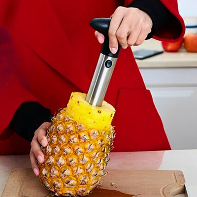 Personne en train d'éplucheur un ananas avec un éplucheur prévu à cet effet posé sur une planche à découper en bois. On voit la moitié de l'ananas sortie de la peau de l'ananas avec une barre métallique.