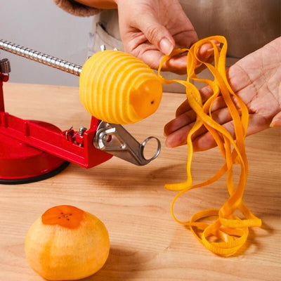 Personne en train d'éplucher un fruit à l'aide d'un éplucheur pomme, l'appareil est posé sur une table, on voit le fruit en train d'être épluchée et coupé, la personne tient les épluchures dans ses mains, en premier plan un fruit déjà épluché.