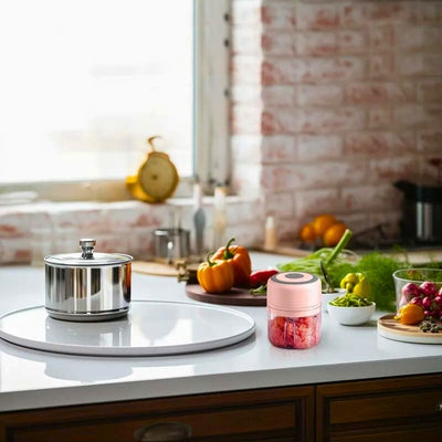 Hachoir électrique rose posé sur un bar de cuisine à côté d'une casserole et de légumes avec en fond une fenêtre et un mur en pierre