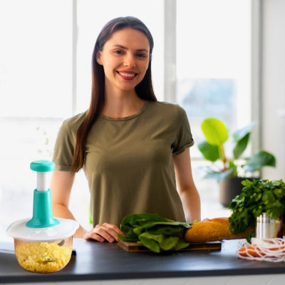 Femme souriante dans sa cuisine devant des légumes posé sur la table, à côté un hachoir manuel rempli d'un légume haché.