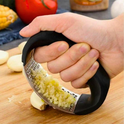 Presse ail tenu par une personne et qui écrase une gousse d'ail sur une planche à découper en bois, on voit également de l'ail déjà pressé sur l'appareil. En arrière-plan, des gousses d'ail et des légumes.