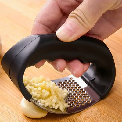 Presse ail tenu par une personne et qui écrase une gousse d'ail sur une planche à découper en bois, on voit également de l'ail déjà pressé sur l'appareil.